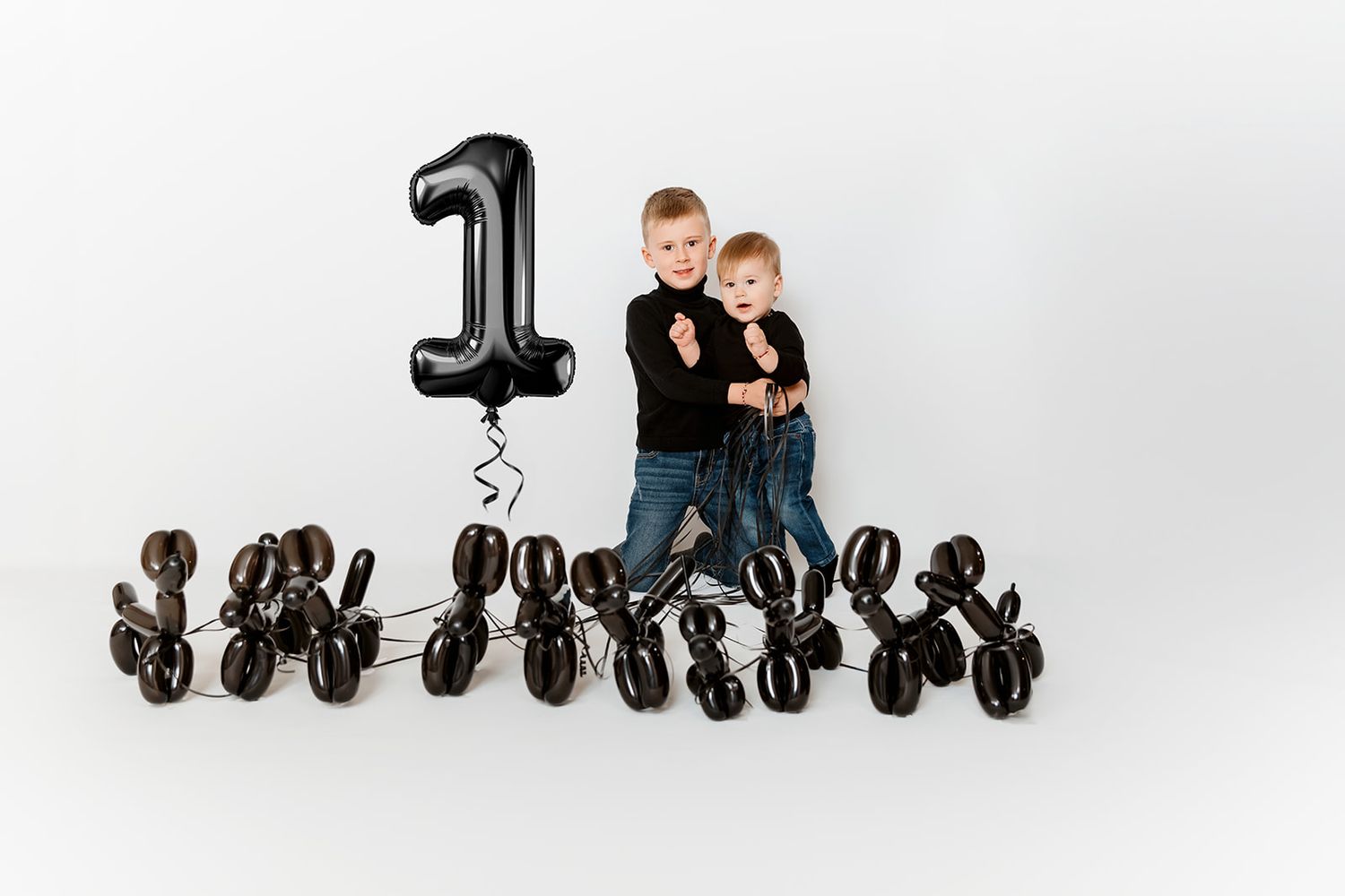 Two boys in black outfits pose with black number one balloon and black balloon dogs for birthday.