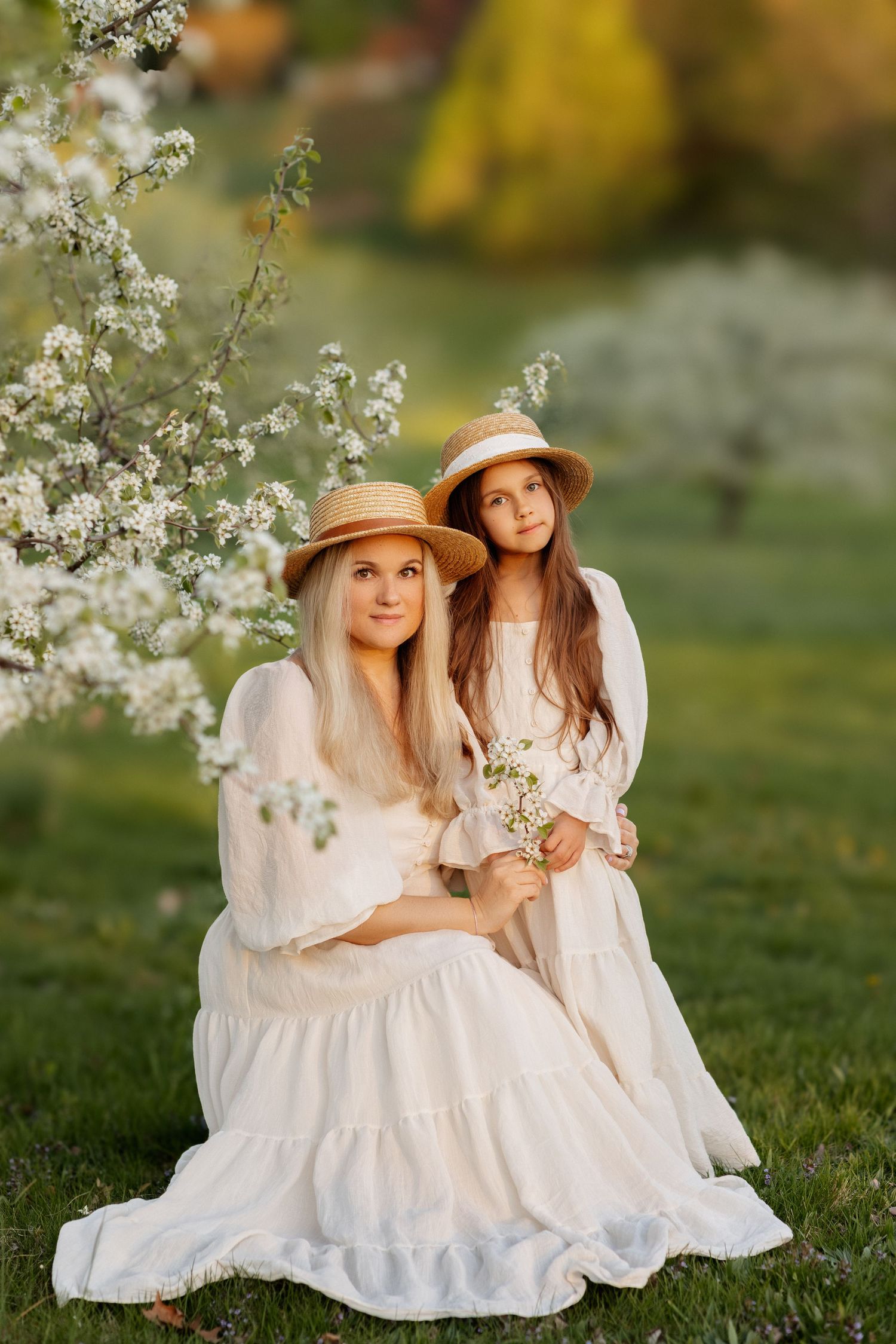 Mother and daughter in white dresses and straw hats pose near blooming tree in sunny garden.