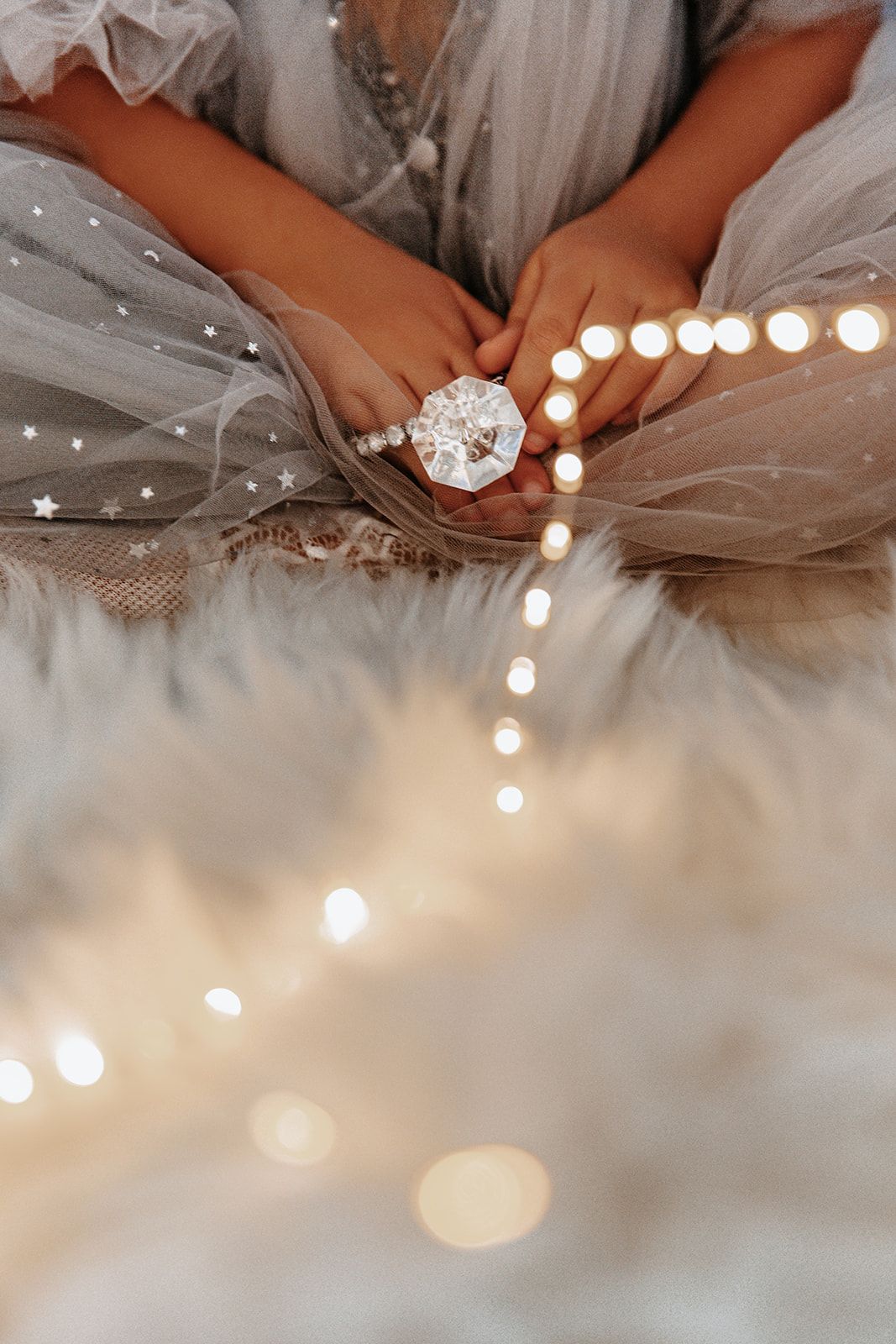 Child in tulle dress holds a sparkling crystal ornament surrounded by glowing fairy lights on a white fur rug.