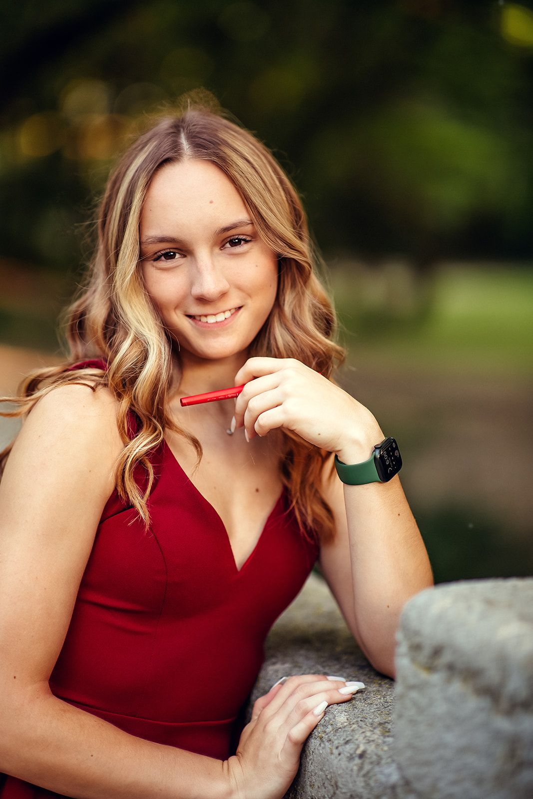 Smiling young woman in red dress holds a red pencil while posing outdoors in a park.