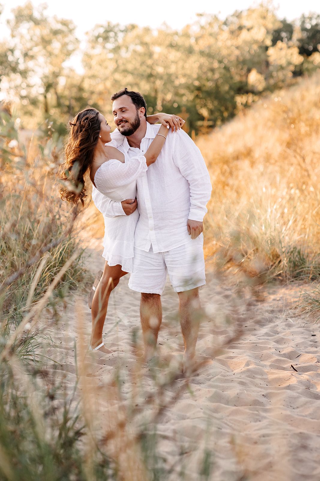 Couple in matching white outfits embracing on a sandy path surrounded by golden grass at sunset.
