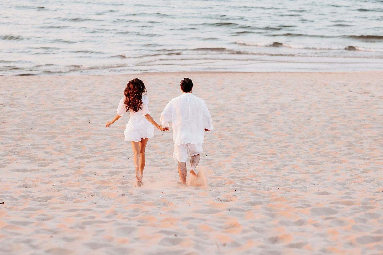 Couple in white outfits running hand in hand on a sandy beach toward the ocean at sunset.