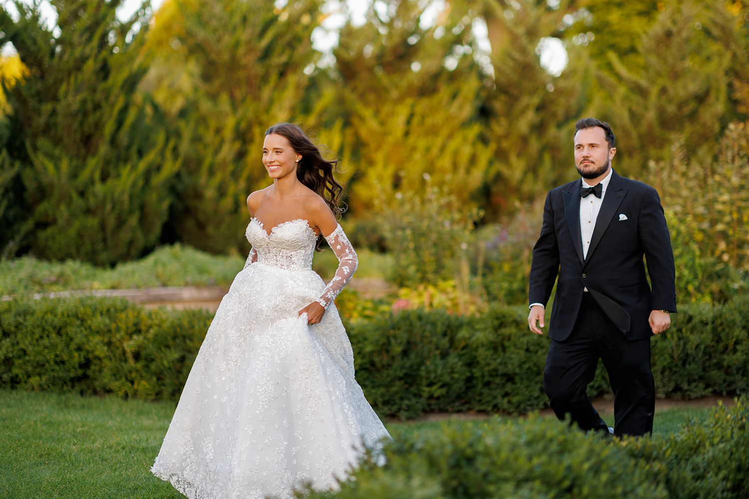 Bride in lace ball gown and groom in black tuxedo walking through lush garden at outdoor wedding ceremony.