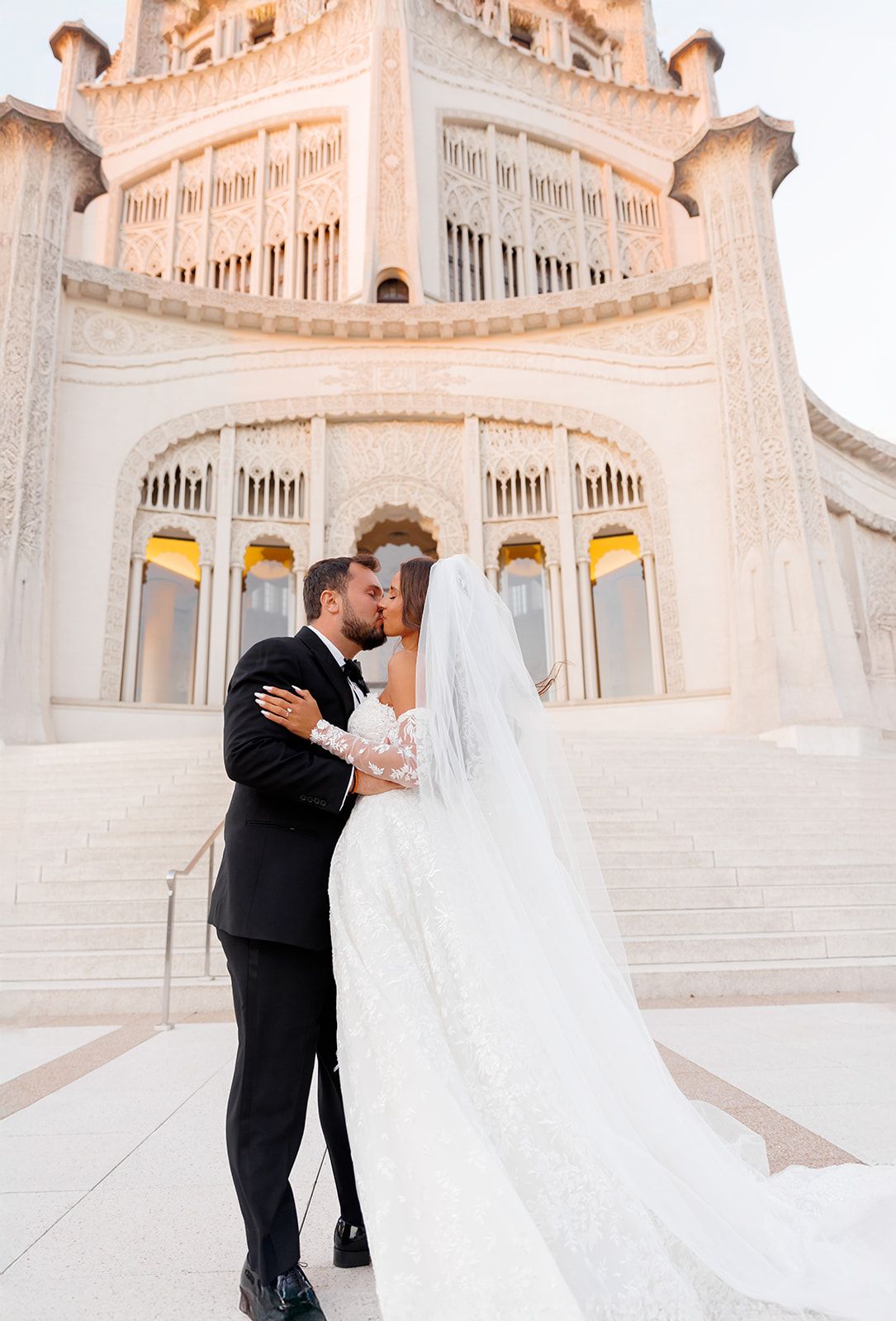 Bride and groom share a romantic kiss in front of an ornate white domed temple at sunset.