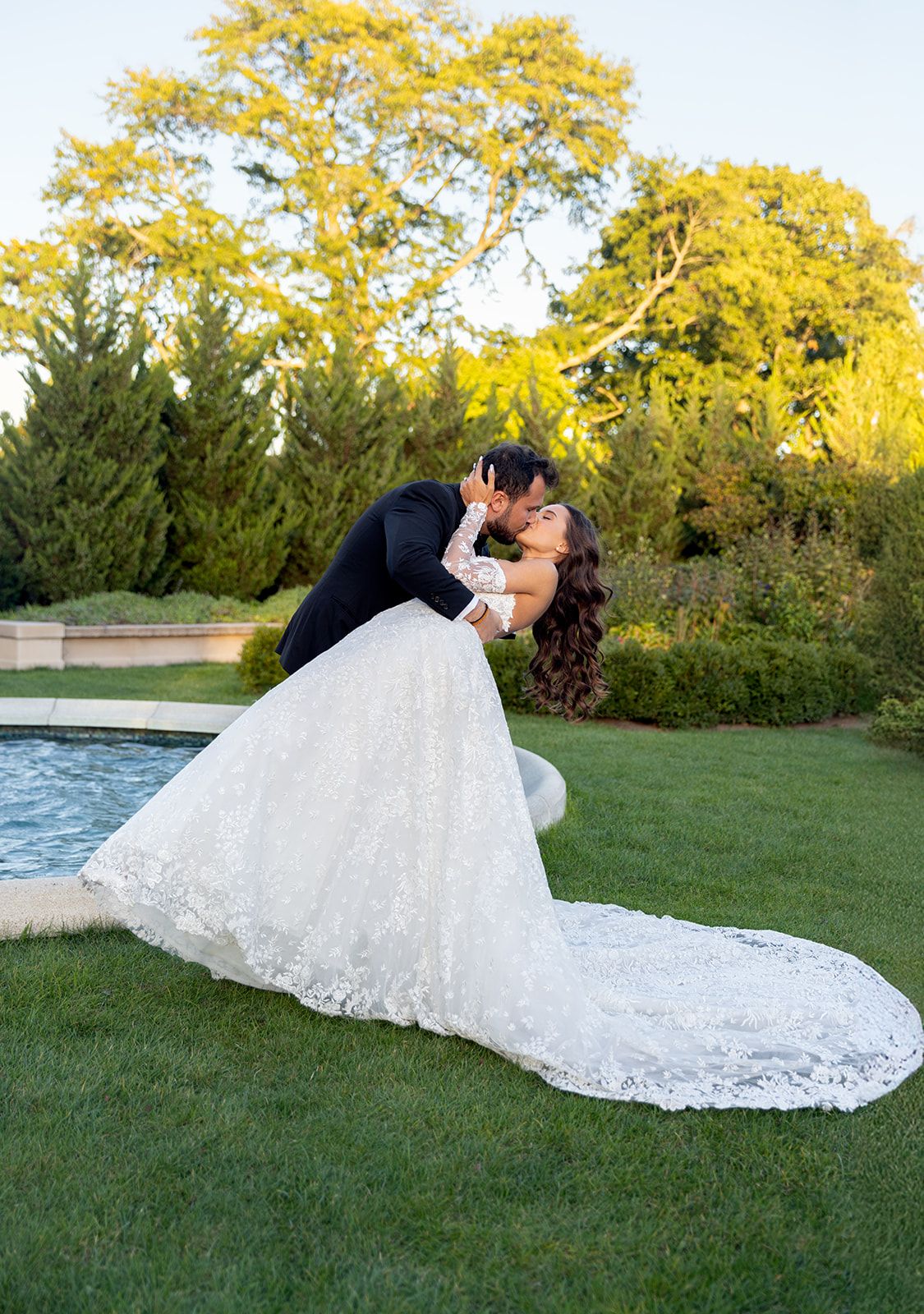 Groom dips bride for a kiss beside a garden pool, her lace gown trailing on the green lawn.