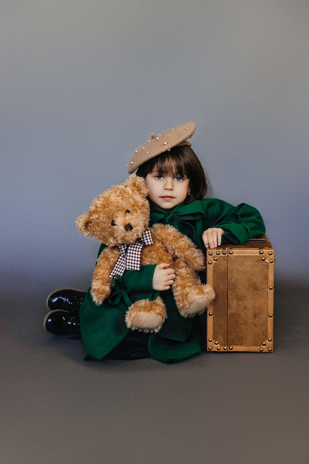 Little girl in green coat and beret hugs teddy bear beside vintage suitcase on gray backdrop.