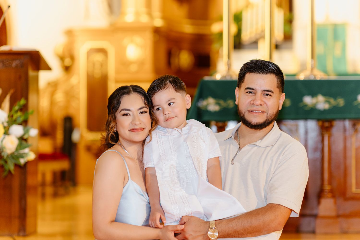 Happy family of three posing in church, parents holding baby in white baptism outfit.