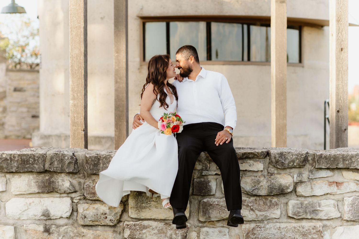 Bride and groom sitting on stone wall, sharing romantic moment with colorful bouquet at outdoor wedding venue.