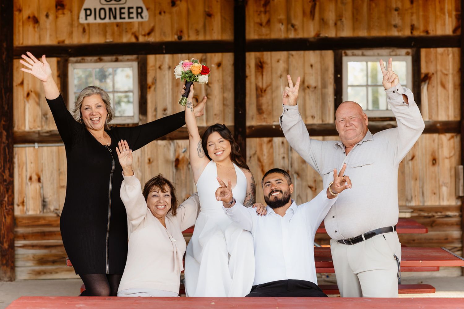 Joyful wedding party celebrates inside Pioneer barn venue, raising hands and bouquet in excitement.