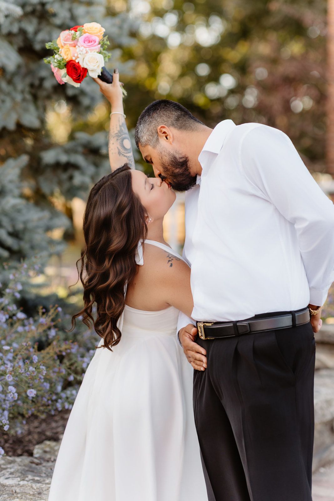 Bride and groom sharing romantic kiss outdoors, bride holding colorful bouquet raised in air on wedding day.