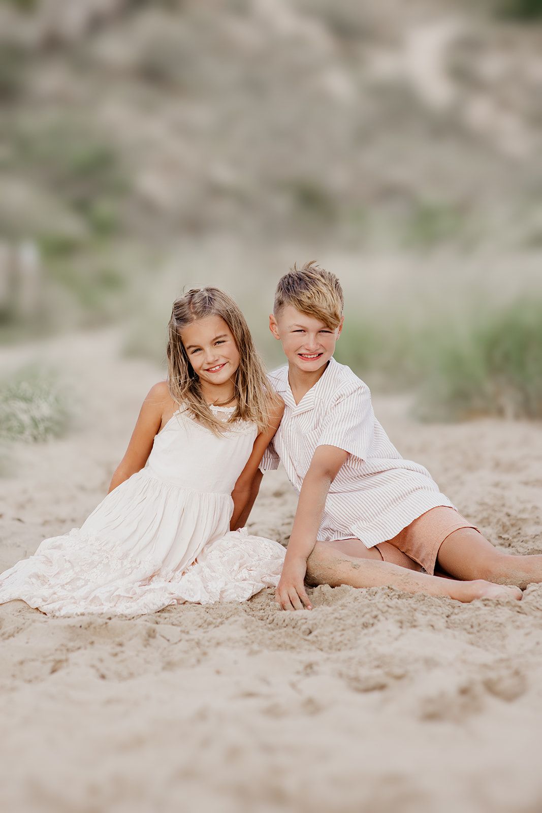 Two smiling children sit together on sandy beach, girl in white dress and boy in light outfit, with soft dunes behind.