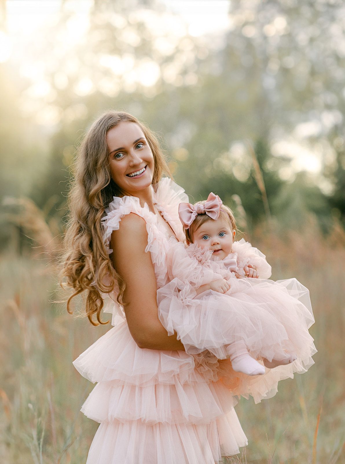 A smiling woman in a pink ruffle dress holds a baby girl in matching pink tulle outfit outdoors.