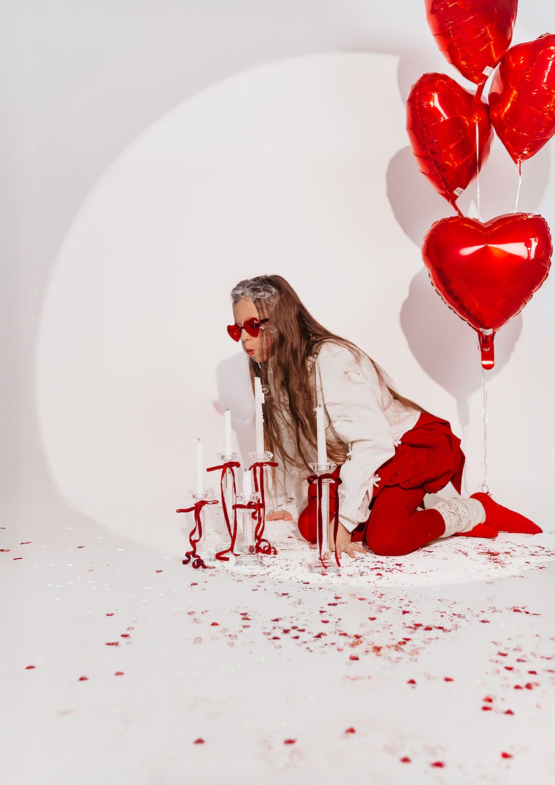 Woman in red pants and heart sunglasses kneels by candles with red heart balloons on a white background.