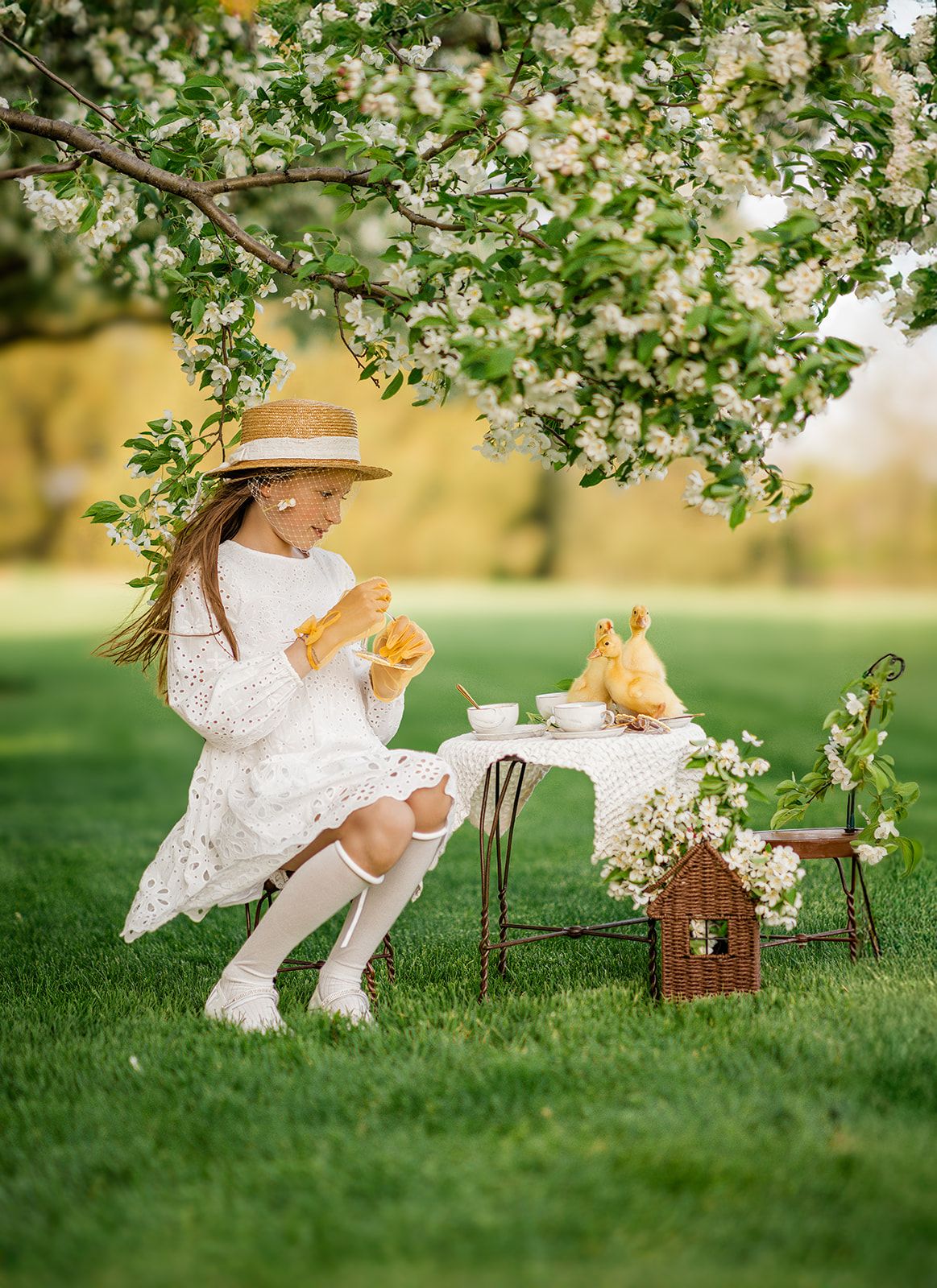 A young girl in a white lace dress and straw hat enjoys a tea party with ducklings under a blossoming tree.