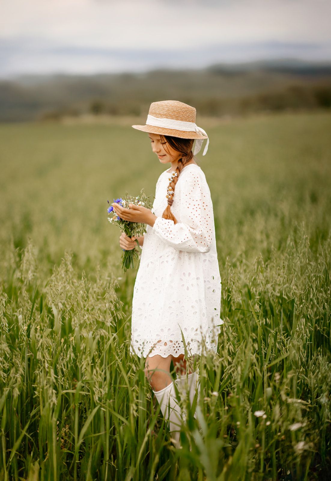 A smiling girl in a white lace dress and straw hat holds wildflowers in a green field.