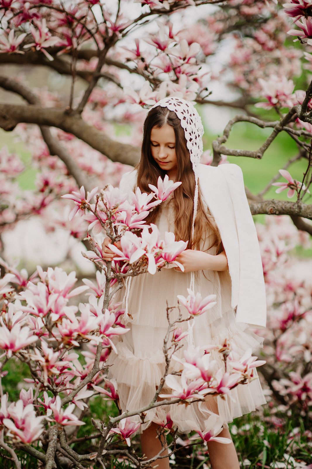 Young girl in white dress and lace bonnet admires pink magnolia flowers in spring garden.