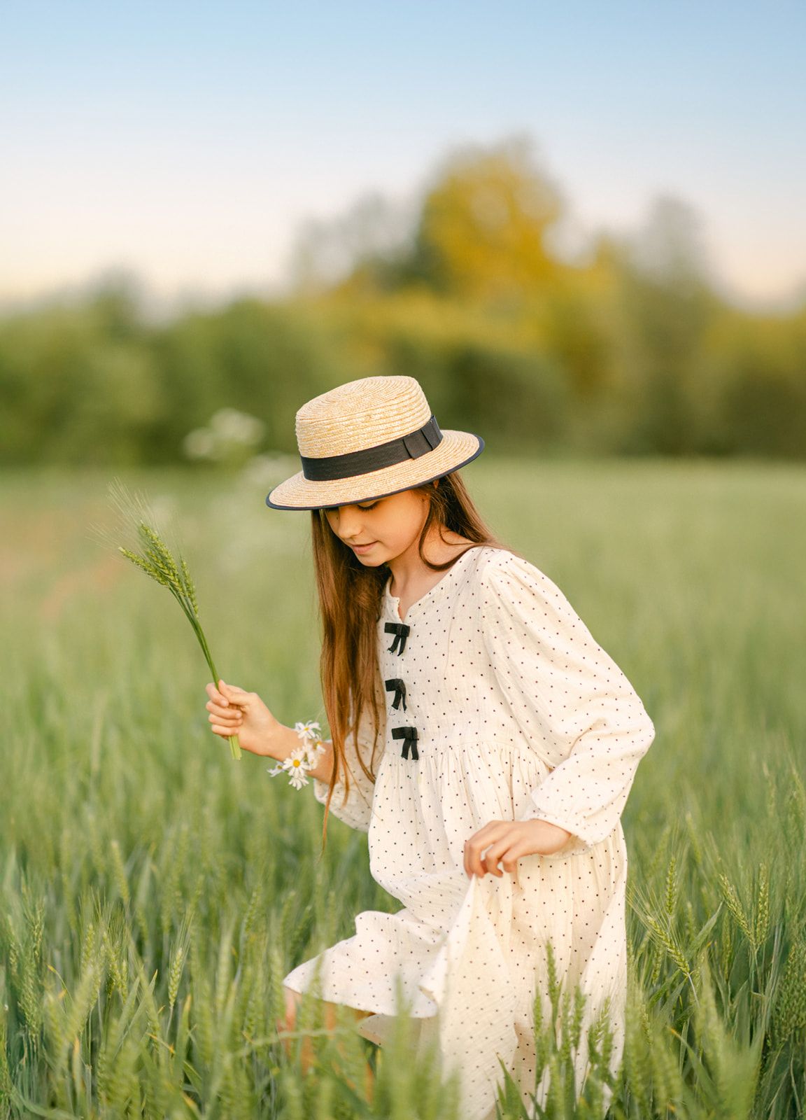Girl in boater hat and white dress picking wildflowers in a lush green wheat field at sunset.