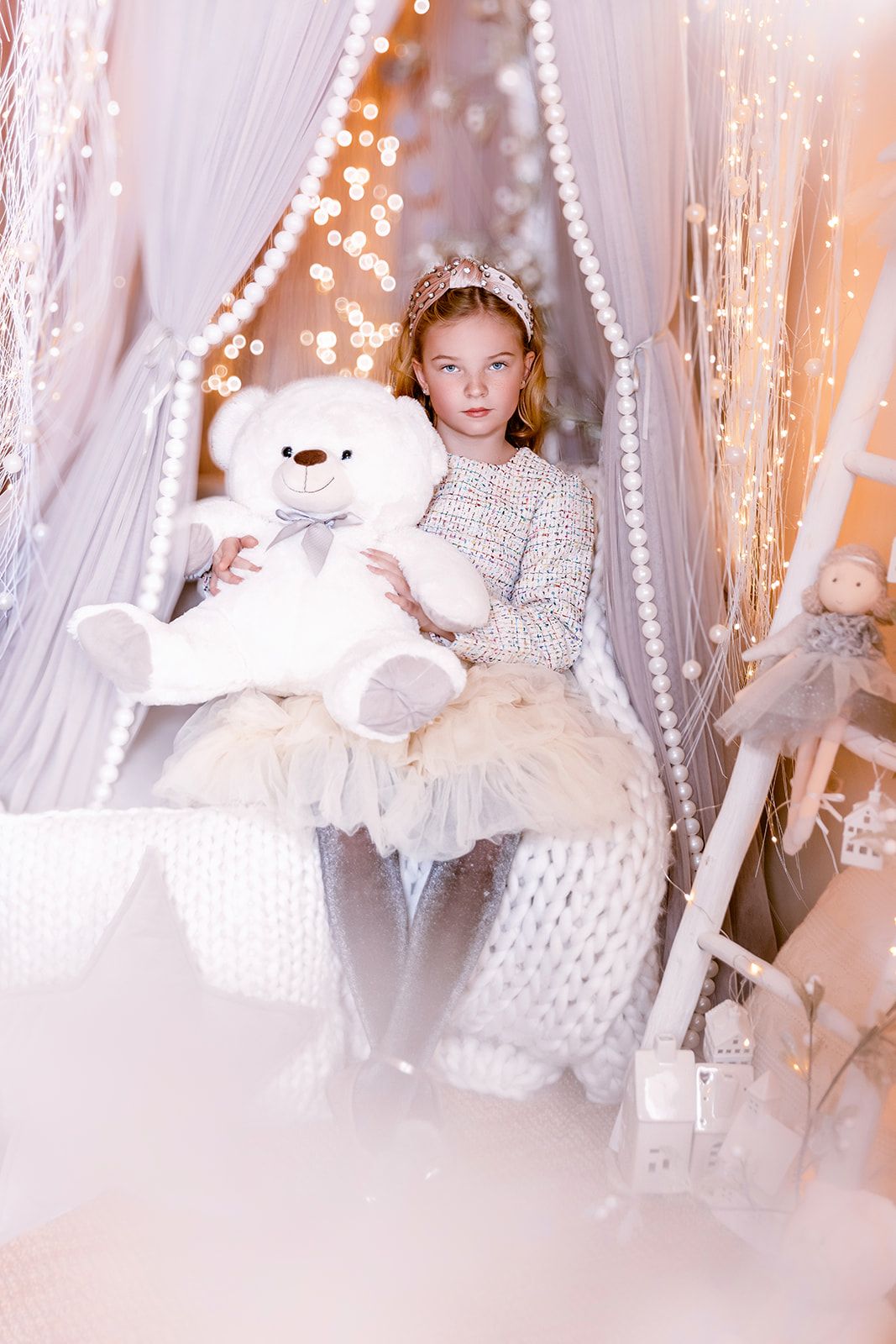 Young girl in white dress holds large white teddy bear, seated in decorated Christmas fairy tale setting.