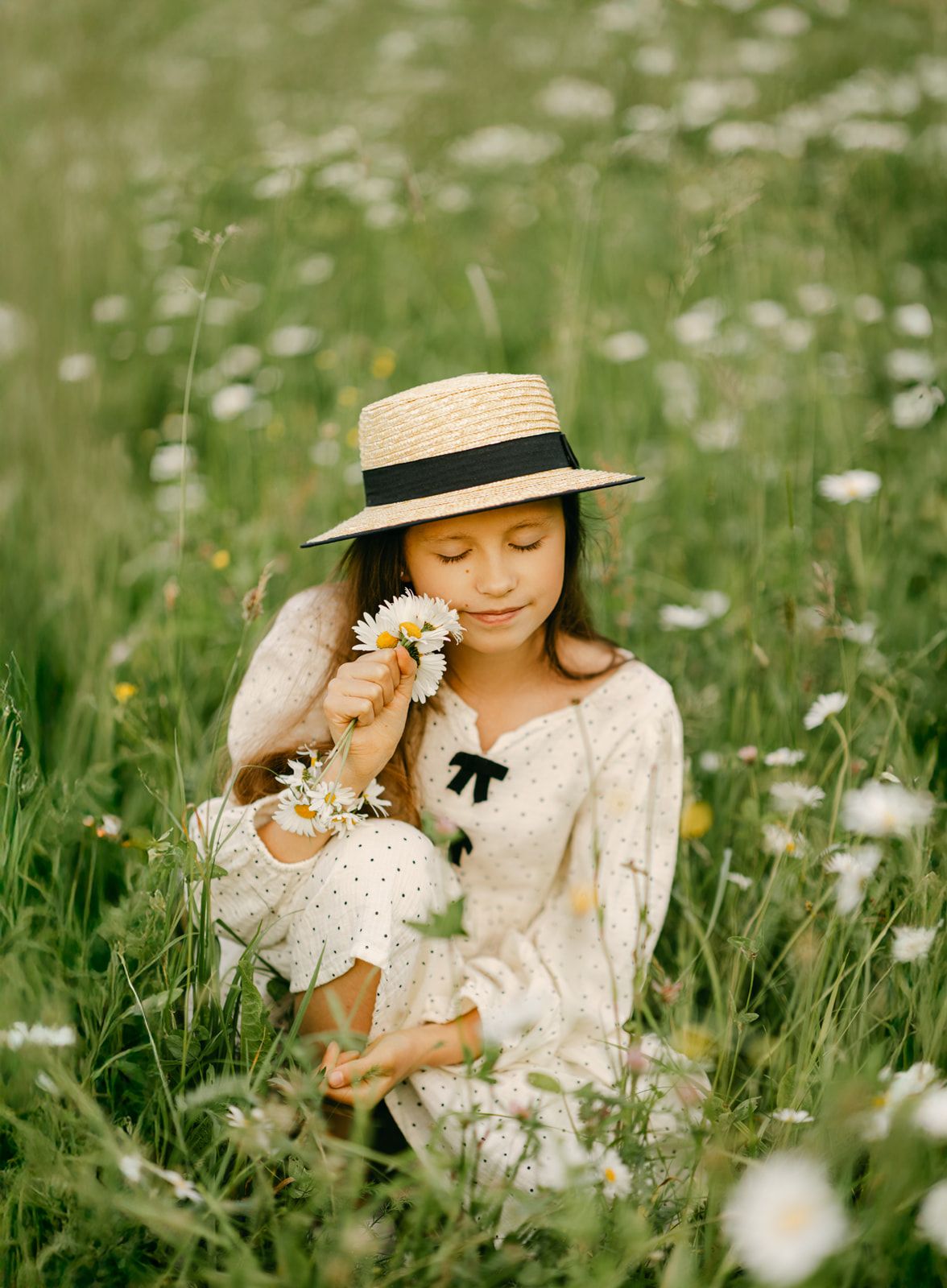 Young girl in straw hat smelling white daisies while sitting in a blooming wildflower meadow.