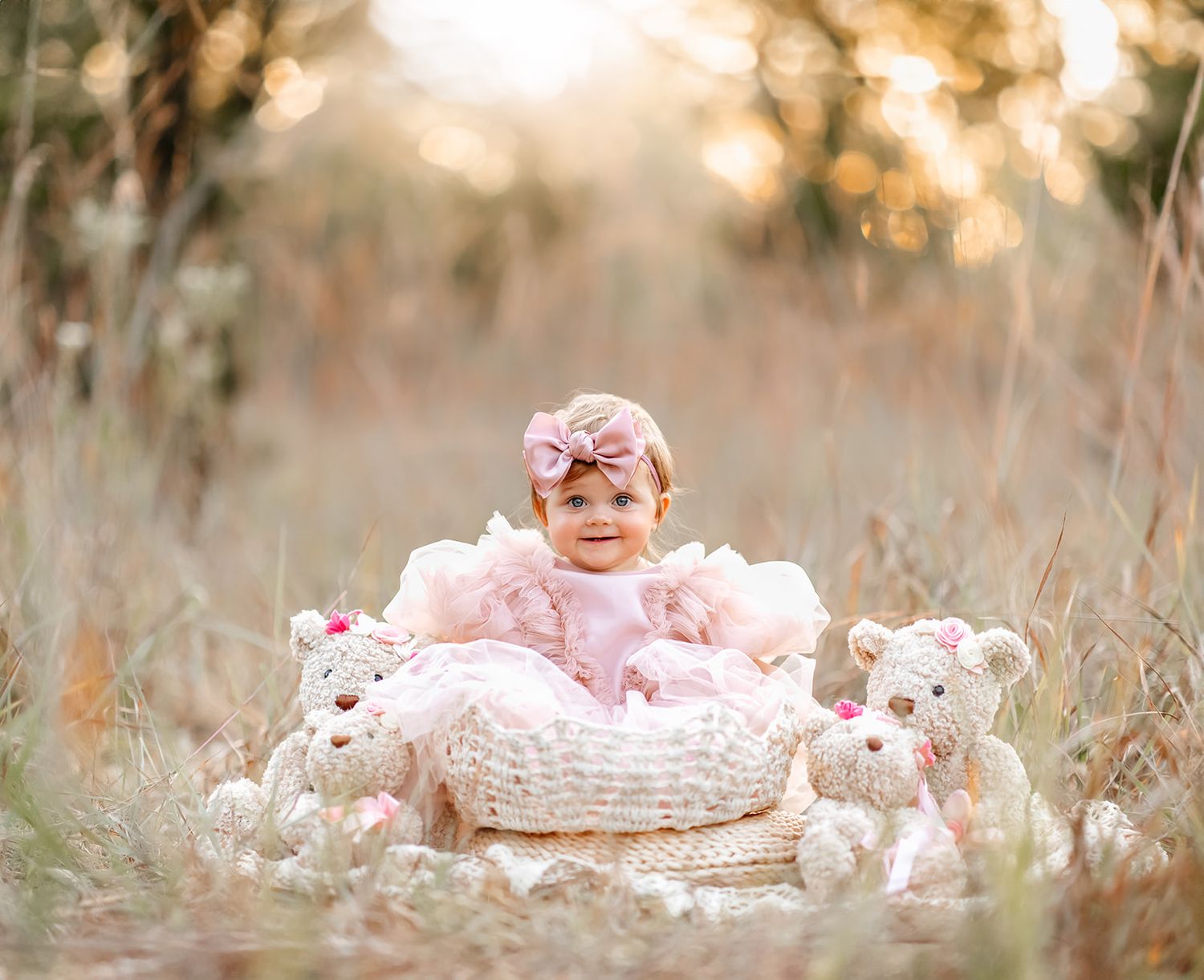 Baby girl in pink dress and bow headband sits in white wicker basket surrounded by teddy bears in a sunlit field.