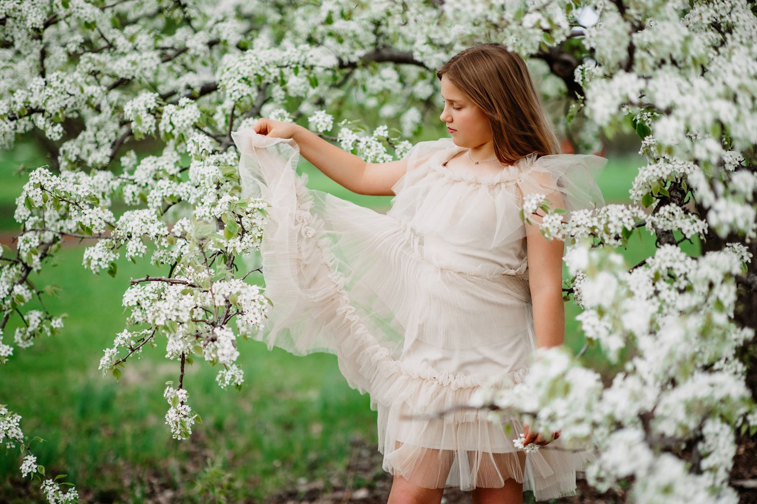 A young girl in a white lace dress poses gracefully among blooming white spring trees in a garden.