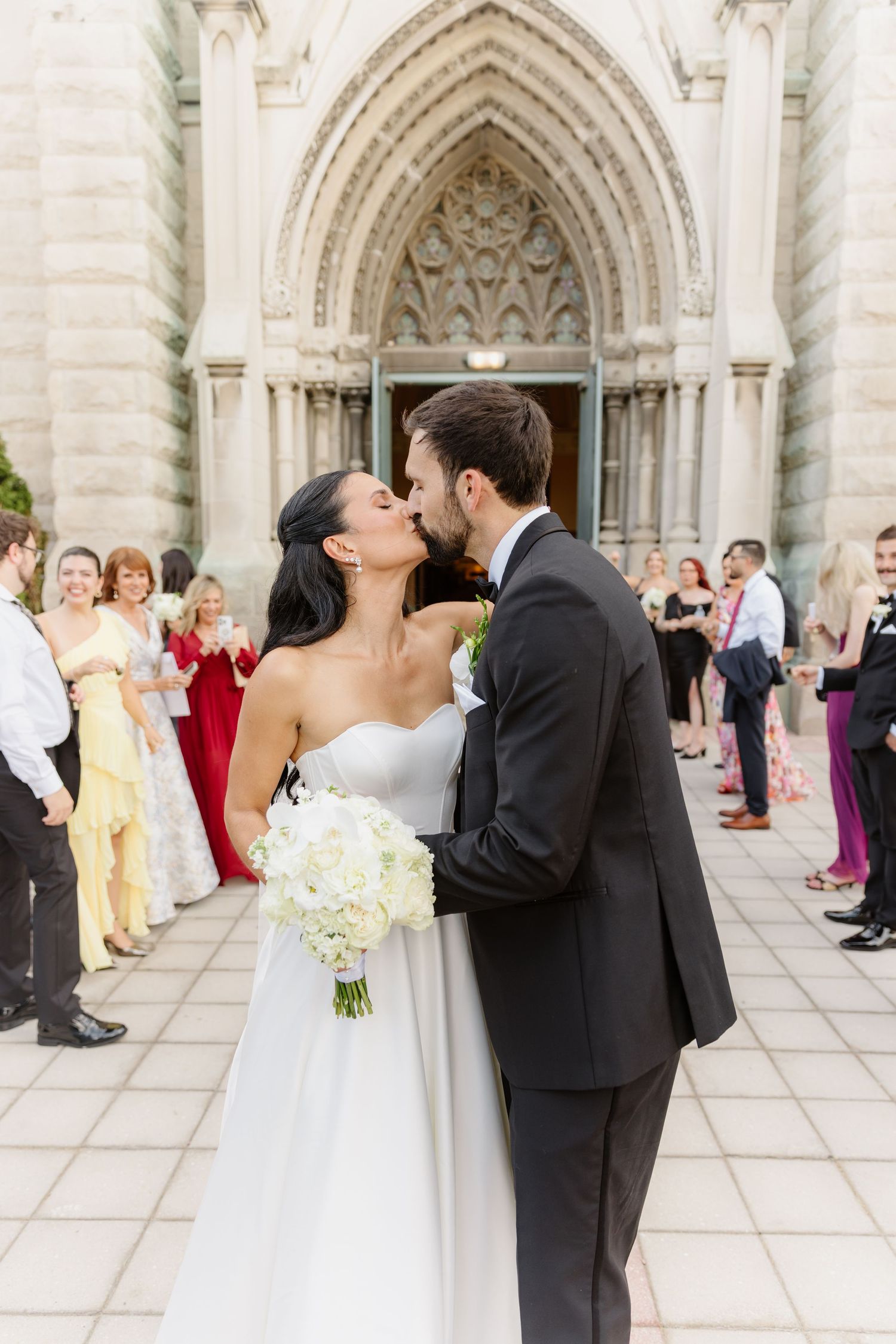 Newlyweds share romantic kiss outside stunning Gothic church entrance after wedding ceremony.