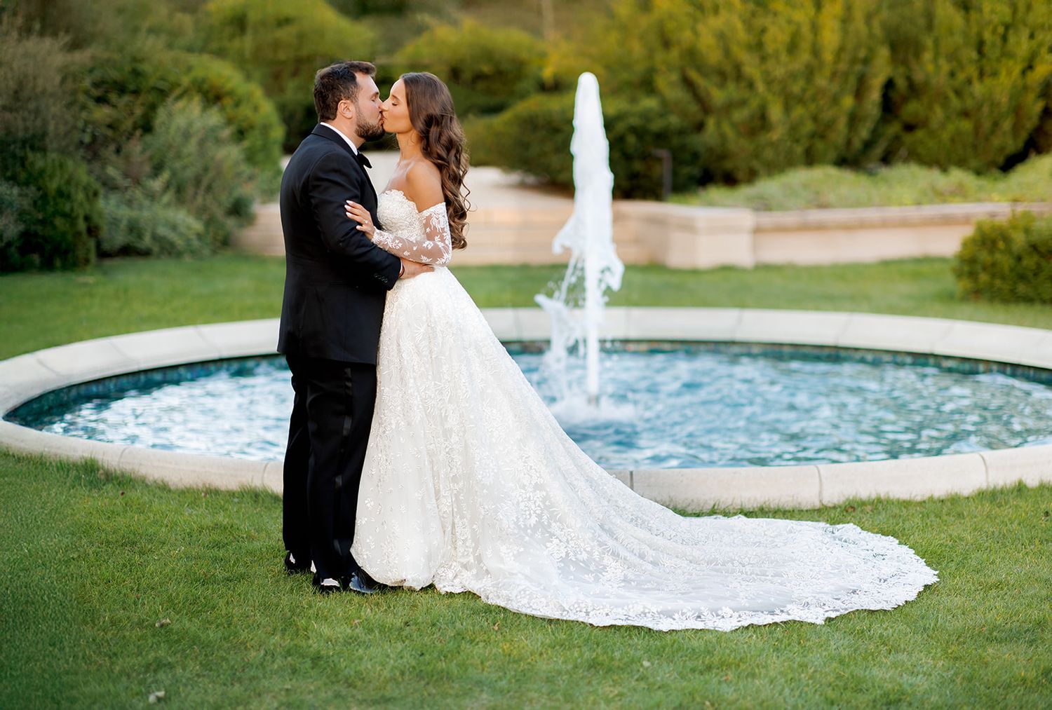 Bride and groom share a romantic kiss in front of a circular fountain in a lush garden wedding venue.