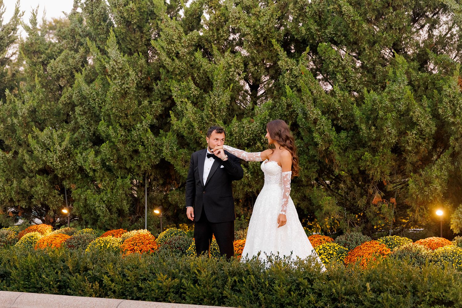 Bride and groom share a romantic dance outdoors, surrounded by evergreen trees and colorful fall chrysanthemums.