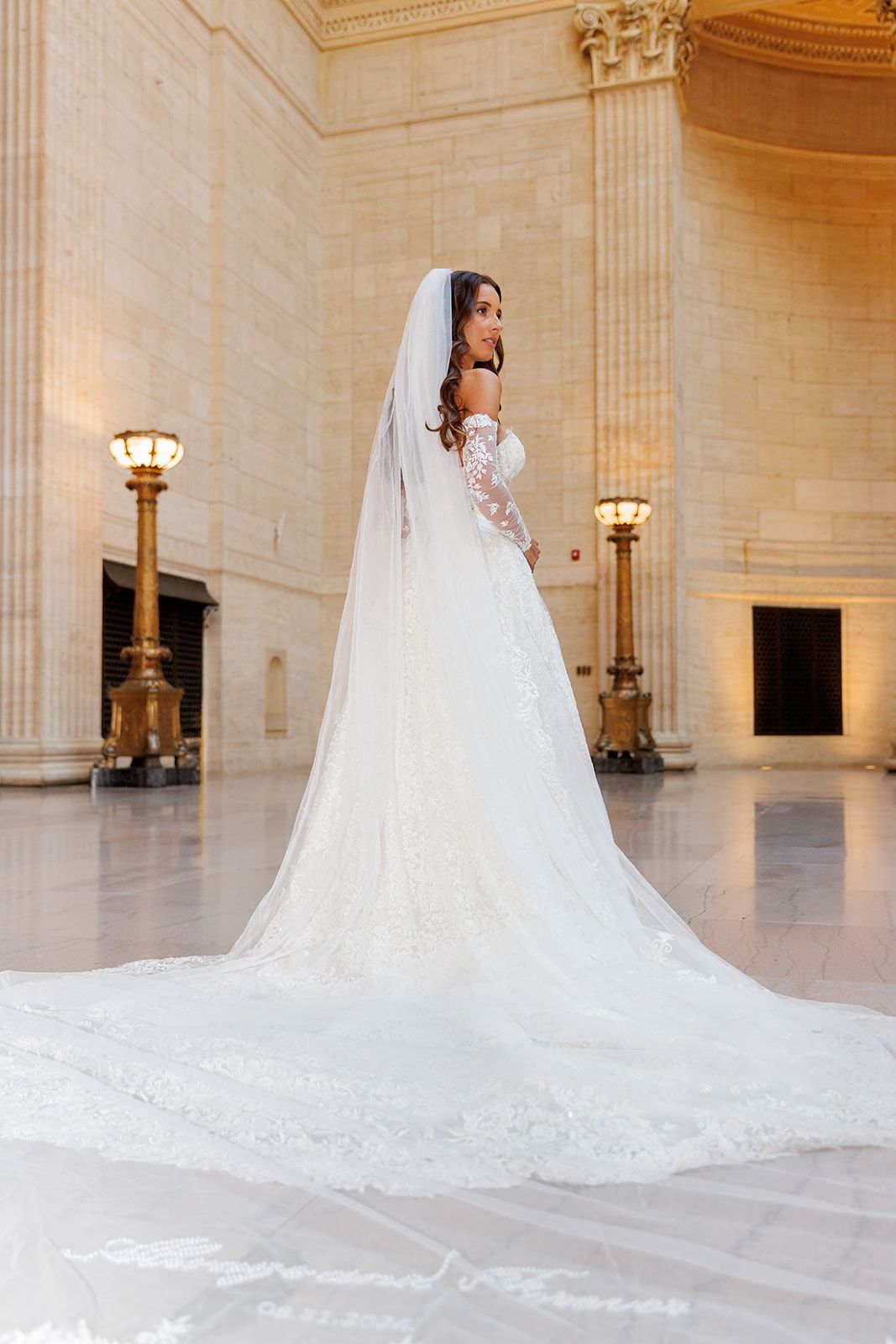 Bride in a stunning off-shoulder lace wedding gown with cathedral veil trains across marble floor of ornate hall.