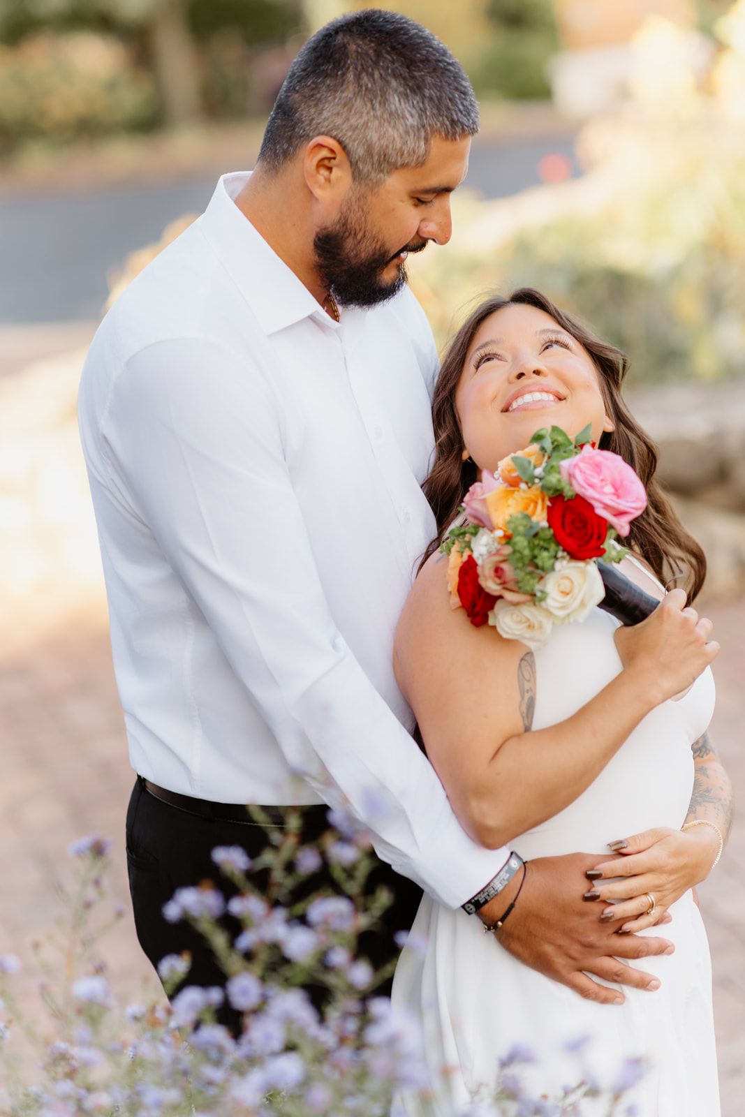 Happy couple embraces outdoors, bride in white dress holding colorful bouquet, groom in white shirt smiling warmly.