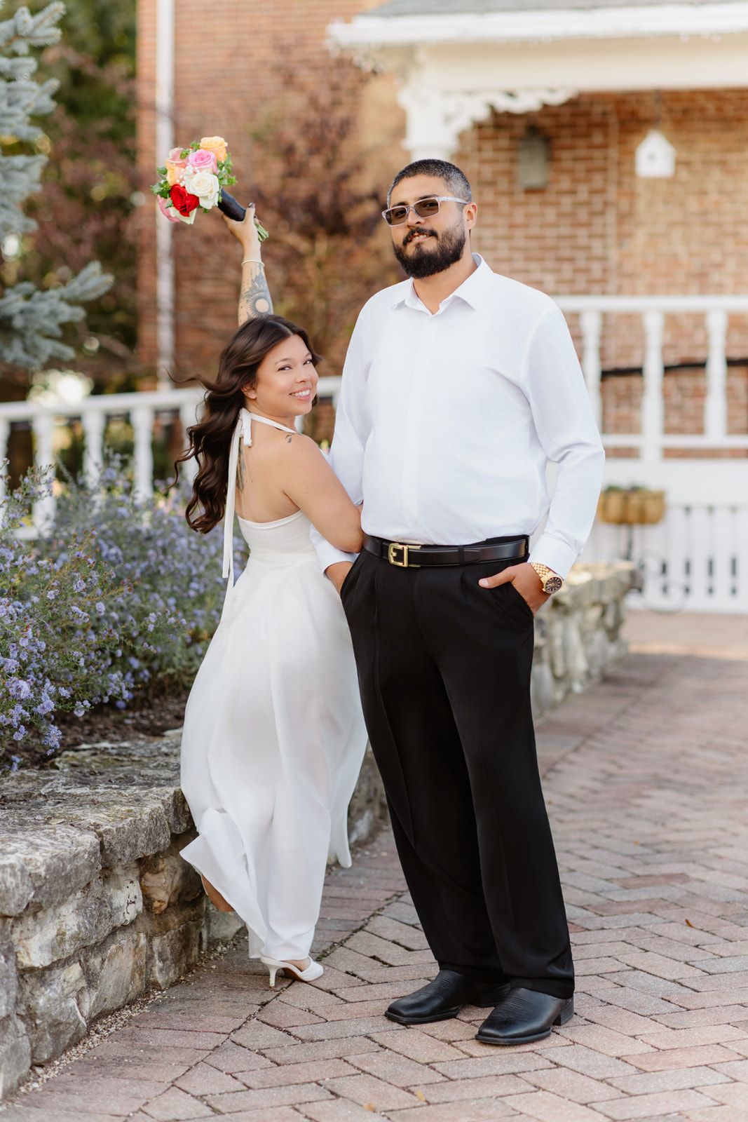 Joyful bride raising colorful bouquet while posing with groom in white shirt on brick pathway.