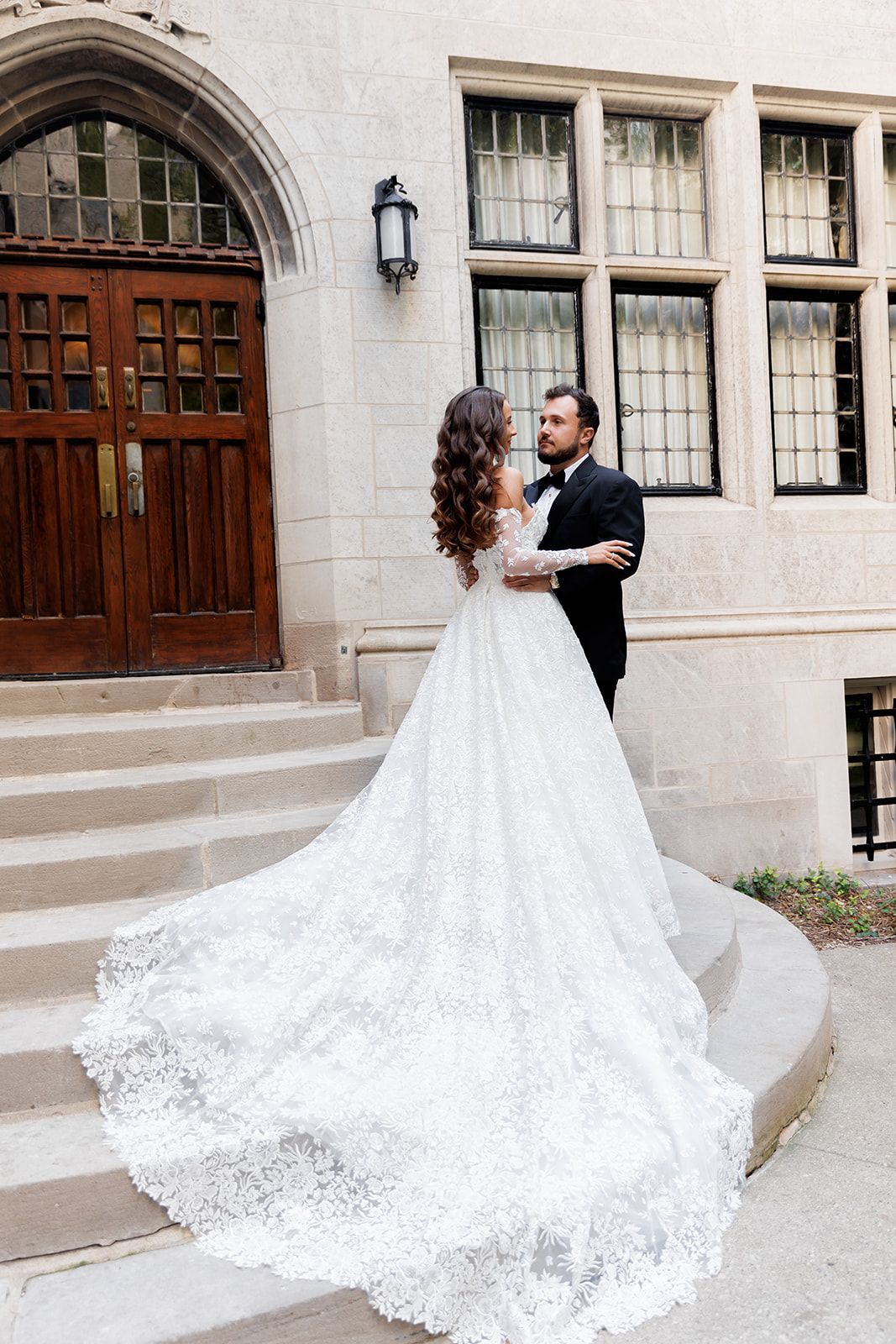 Bride in lace gown with long train stands with groom in tuxedo on stone steps outside elegant building.