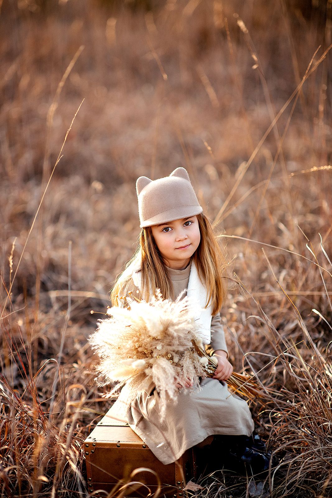 A young girl in a beige cat-ear hat holds dried pampas grass while sitting on a suitcase in a golden field.