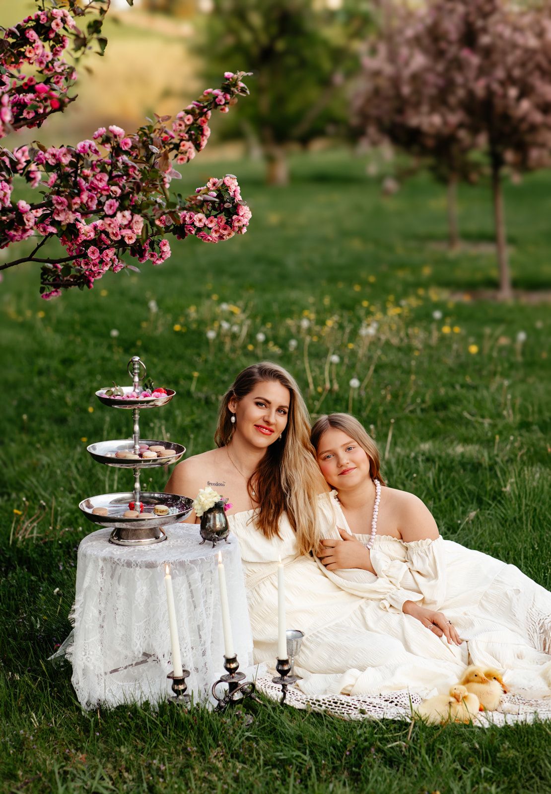 Woman and girl in white lace dresses pose by tiered dessert stand with candles under pink flowering tree outdoors.