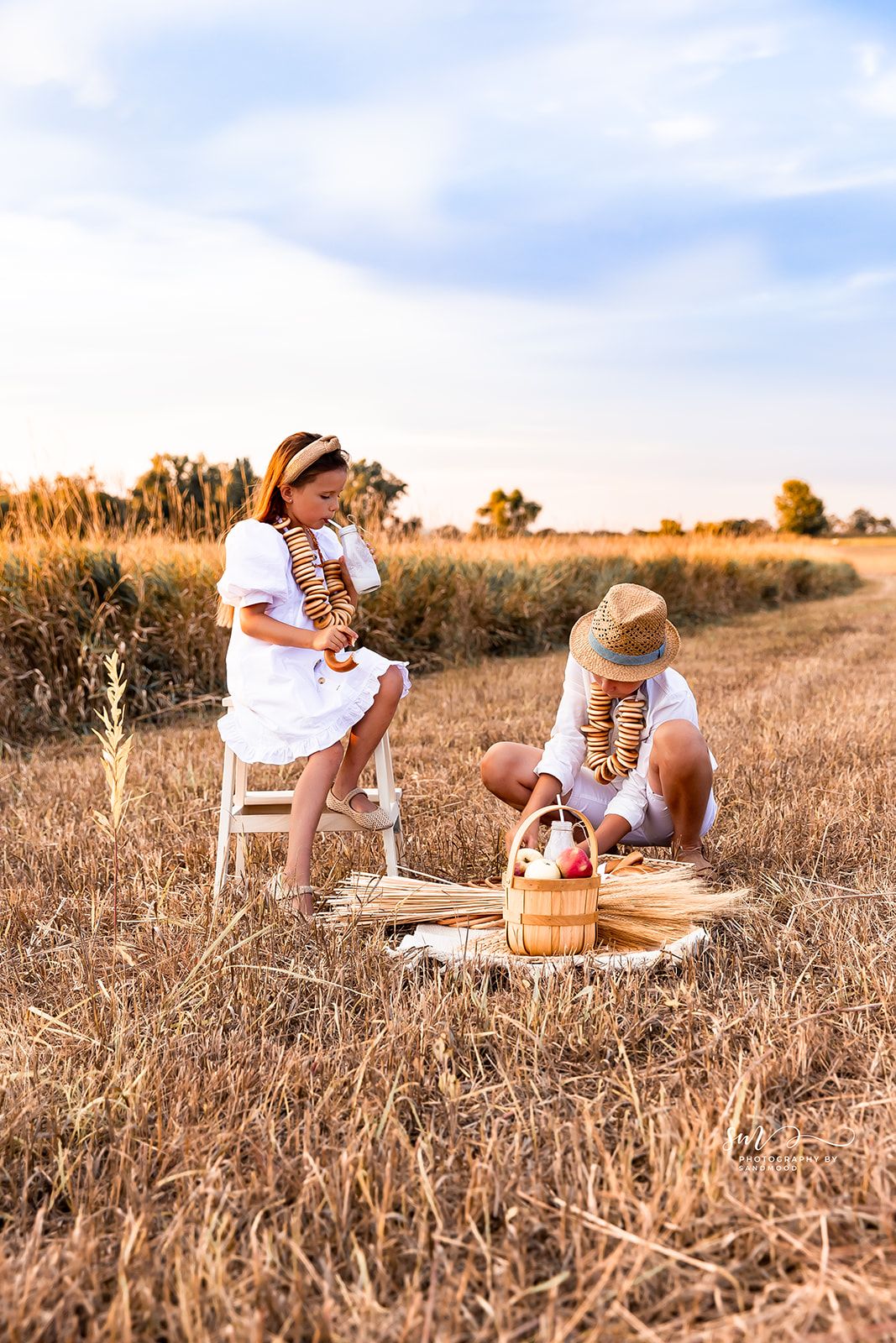 Two children in white dresses enjoy a countryside picnic in a golden wheat field at sunset with a basket of fruit.
