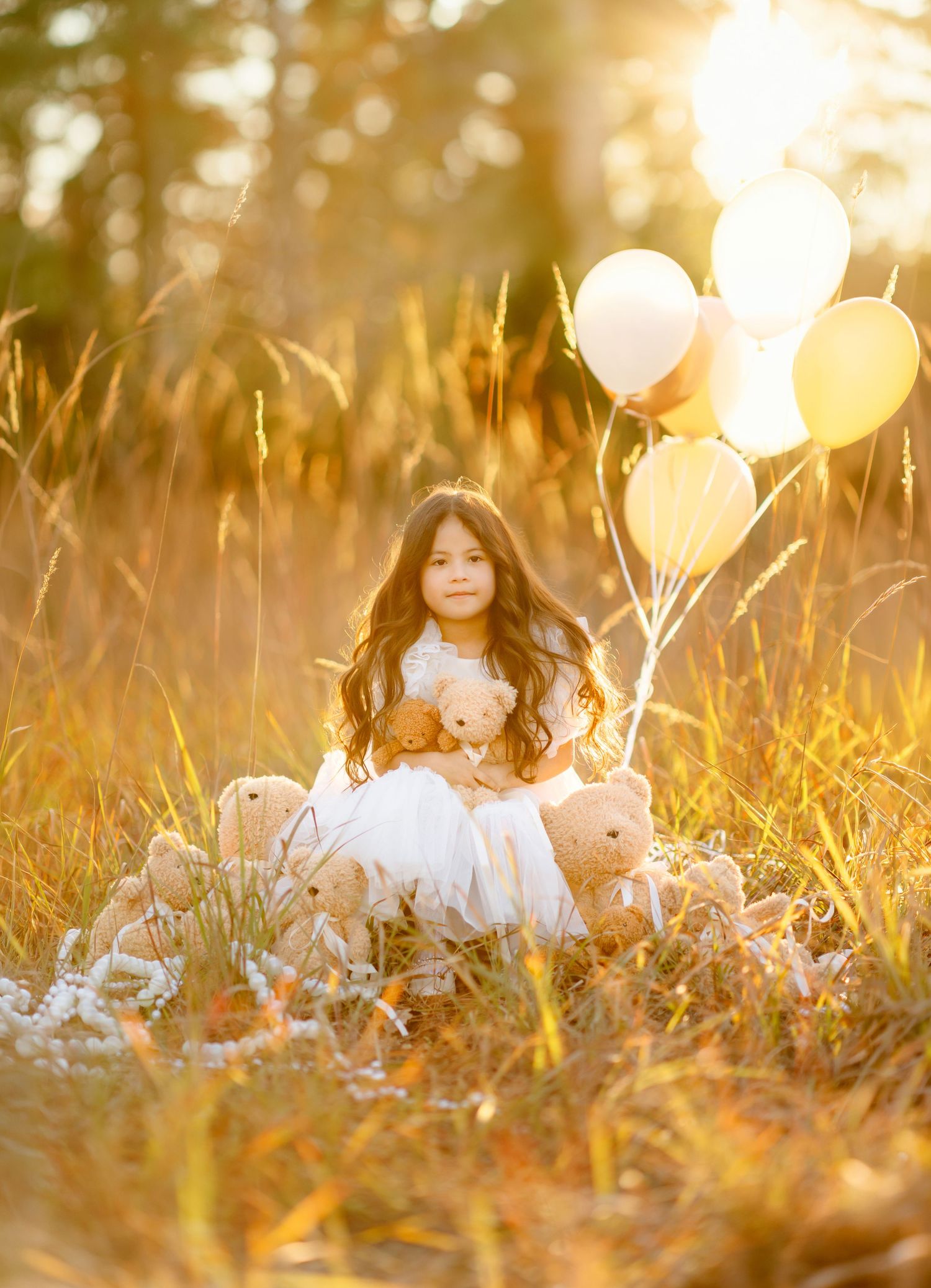 A young girl in a white dress sits in a golden field with teddy bears and balloons in warm sunlight.