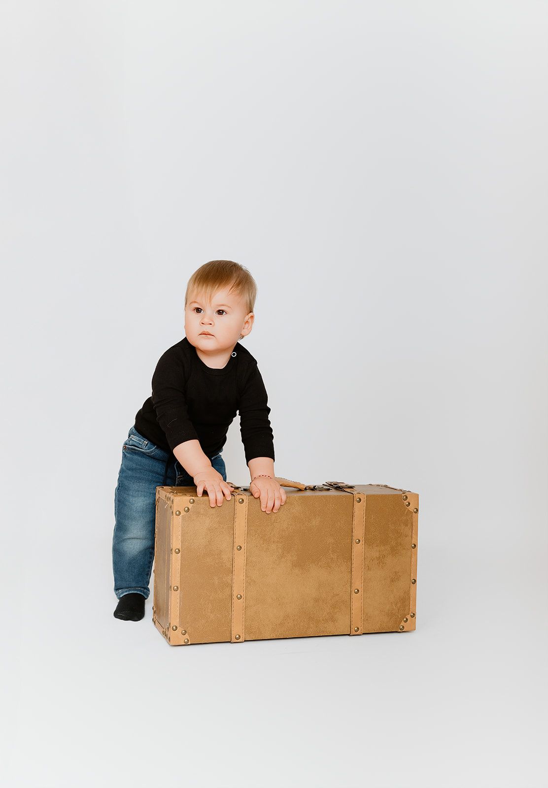 Toddler in black sweater and jeans leaning on a vintage tan suitcase against a white background.