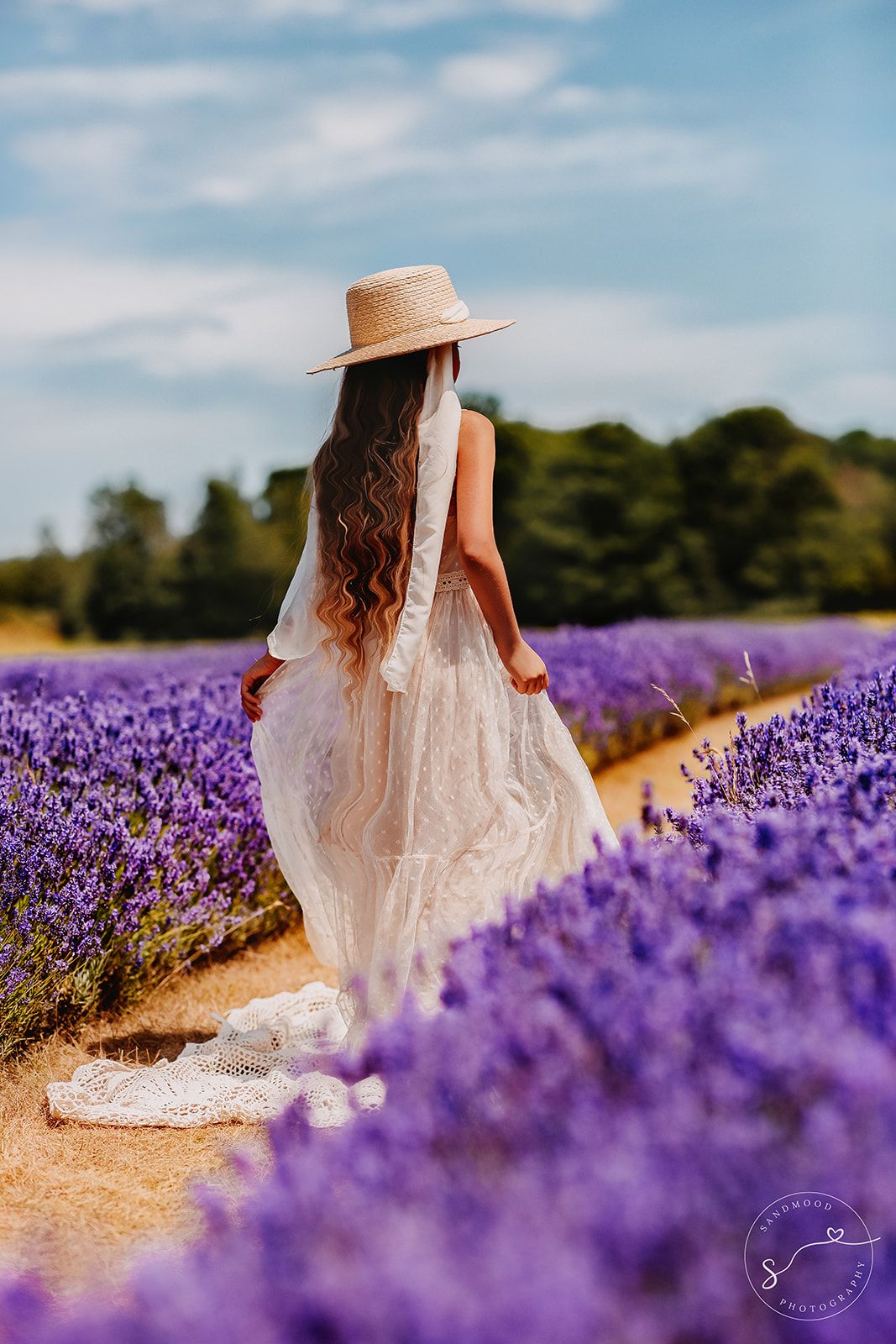 Woman in white lace dress and straw hat walking through vibrant purple lavender field.
