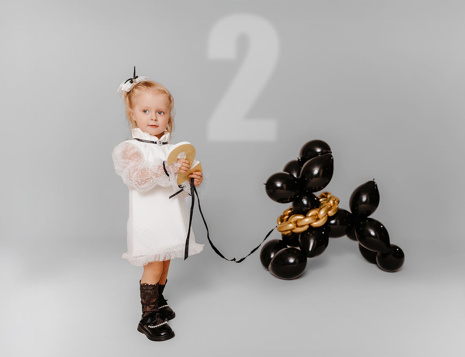 Toddler girl in white dress holding balloon dog leash next to number 2 for second birthday photo.