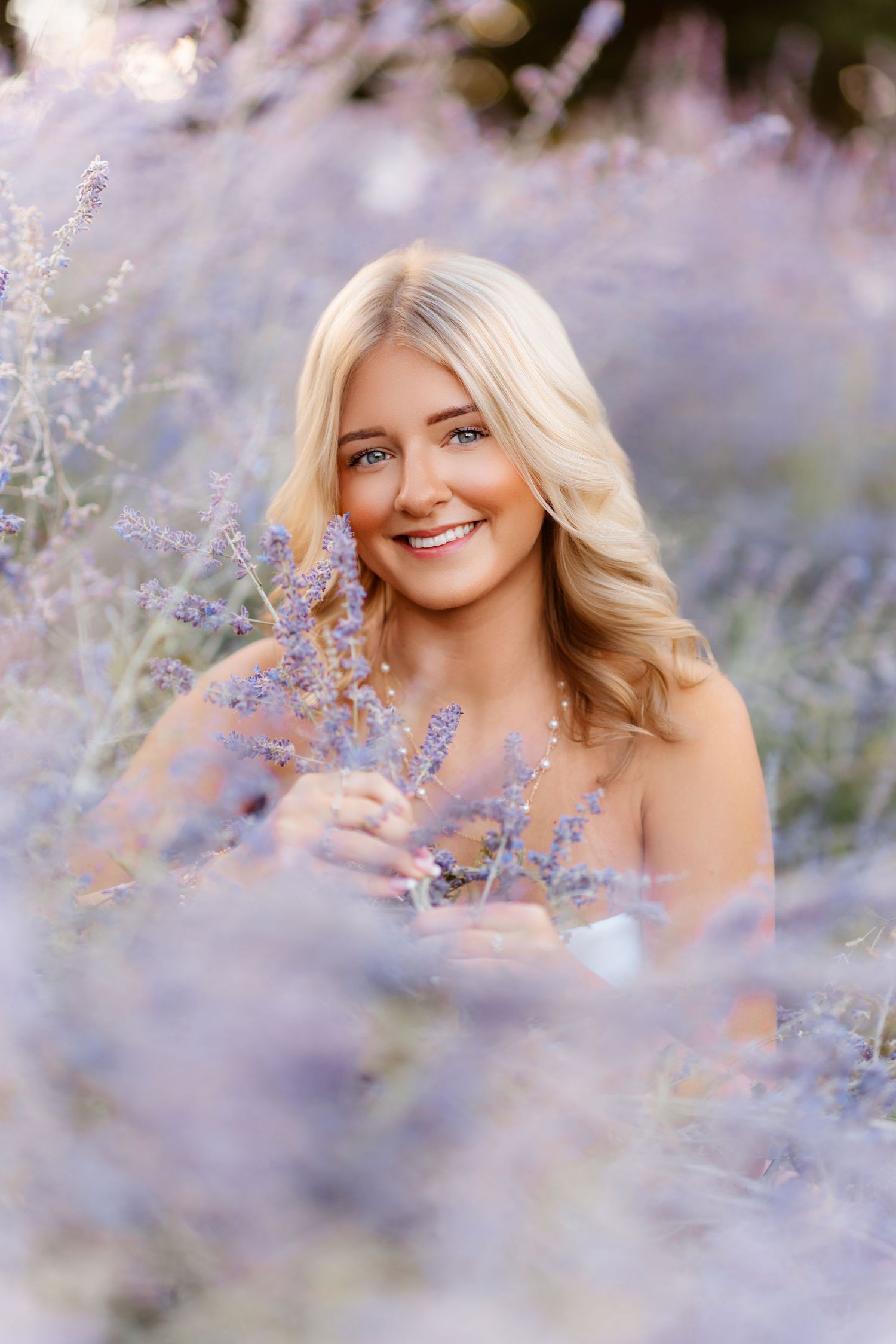 Smiling blonde woman surrounded by blooming purple lavender flowers in soft light.