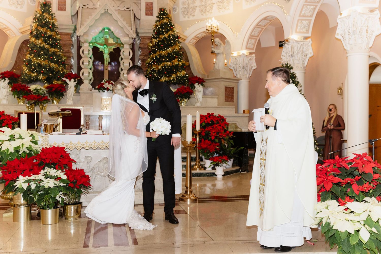 Bride and groom stand before priest at ornate Christmas-decorated church altar during wedding ceremony.