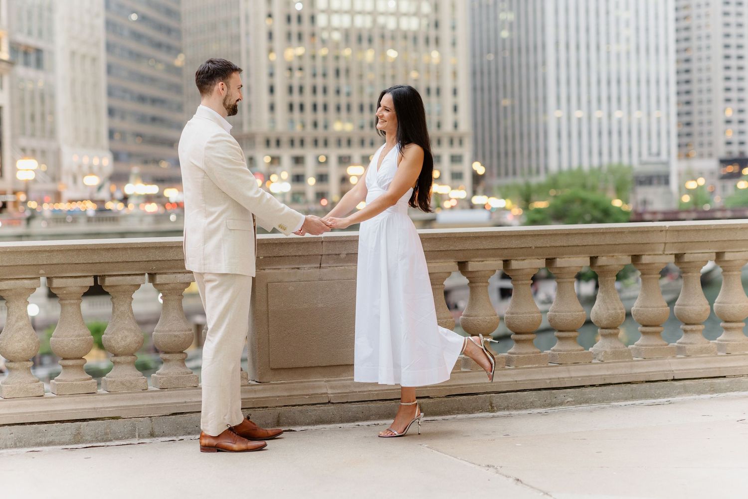 Engaged couple holding hands face-to-face on stone balcony with Chicago skyscrapers in the background.