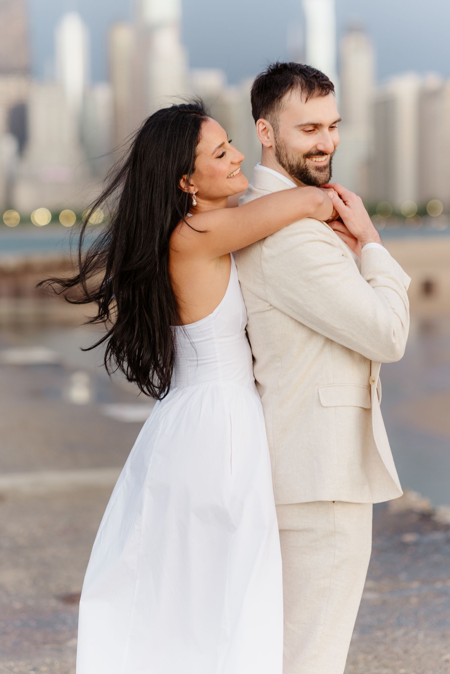 Smiling bride in white dress hugging groom from behind with Chicago skyline and lake in background.