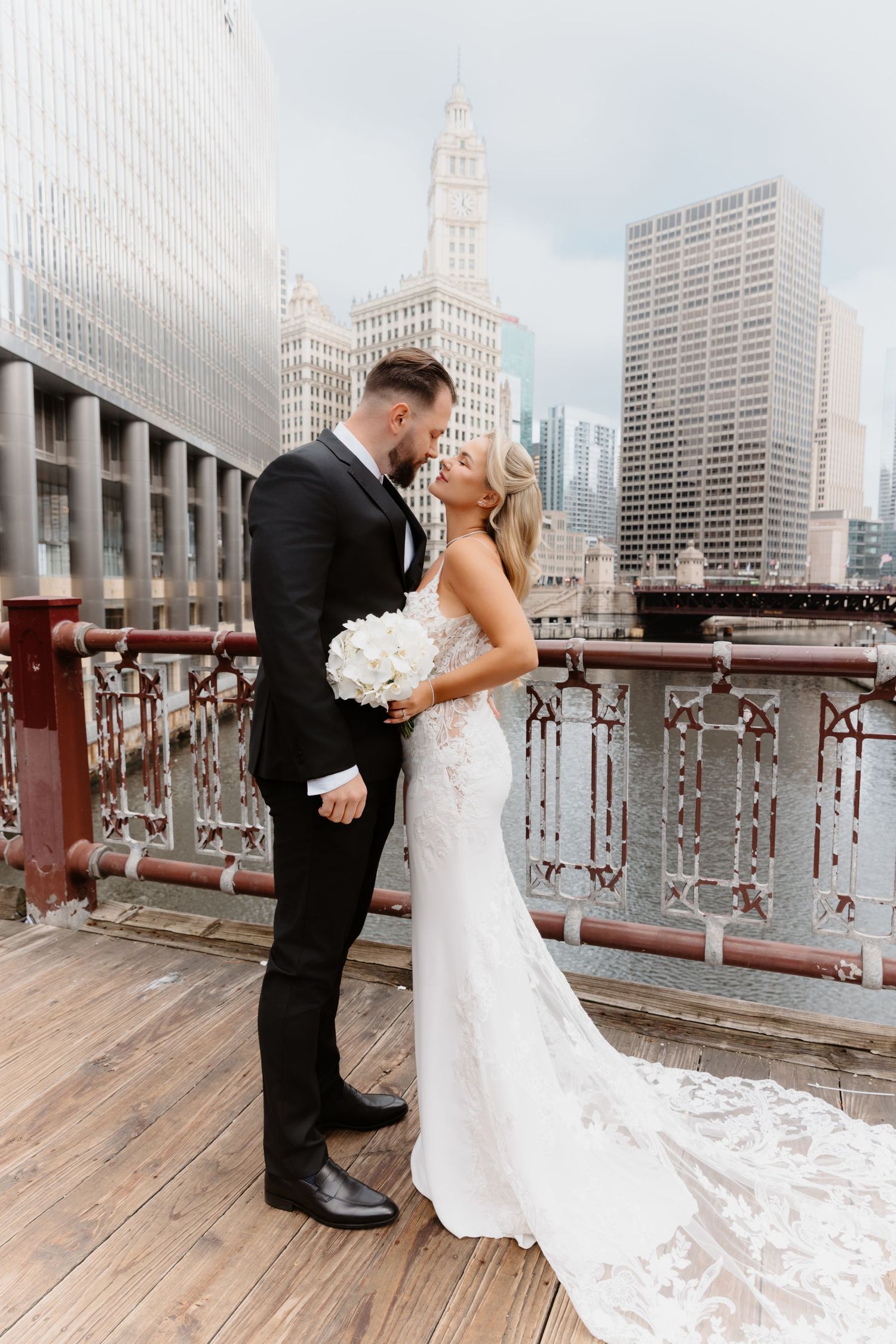 Bride and groom nearly kiss on Chicago river bridge, bride holding white bouquet with city skyline behind.