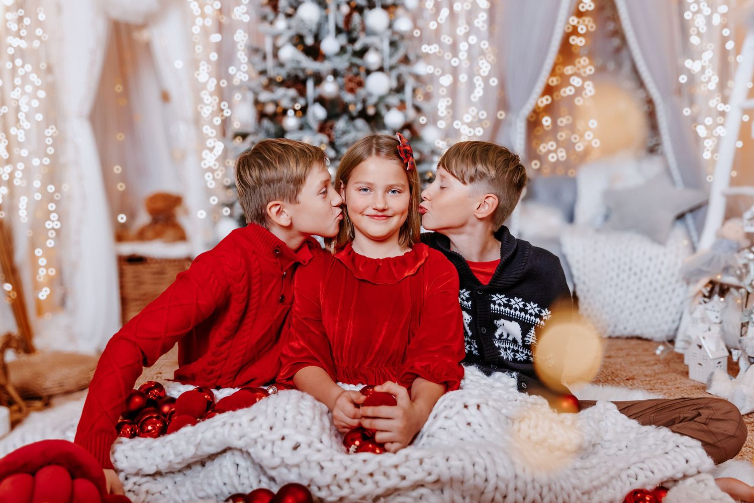 Two boys kiss a girl on the cheeks while sitting together on a blanket near a Christmas tree.