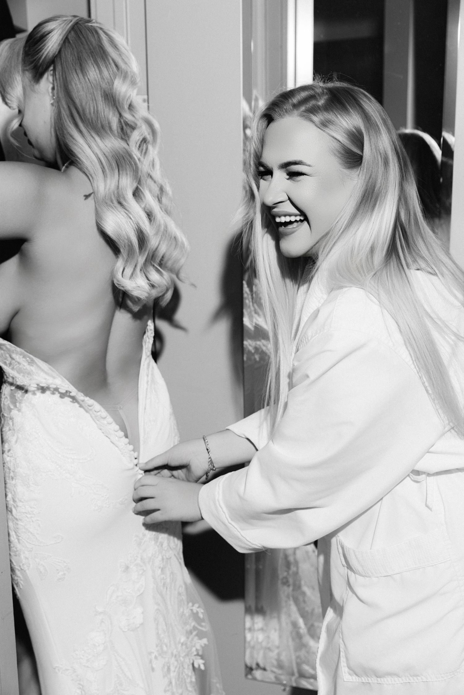 Smiling bridesmaid in white robe buttoning up brides lace wedding dress in black and white bridal preparation photo.