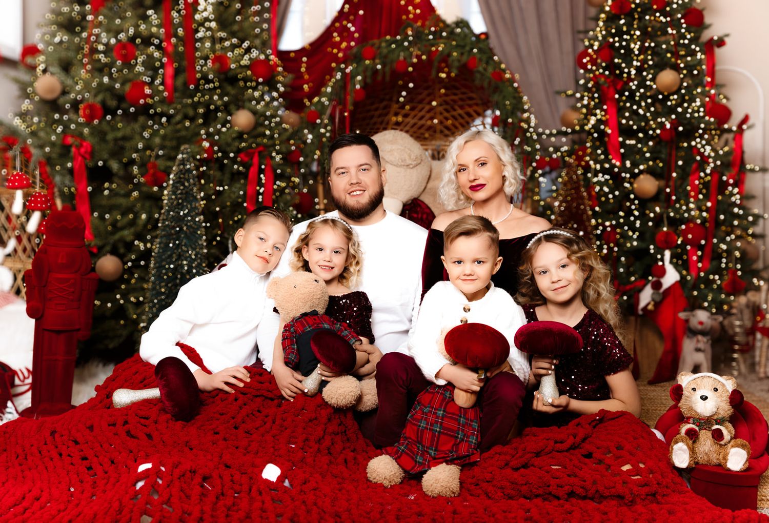 Family of six poses for a festive Christmas photo surrounded by decorated trees and red holiday decor.