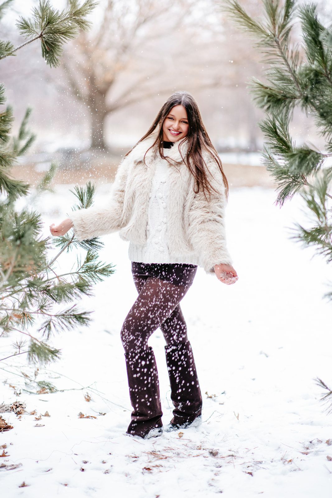 Young woman in white fur coat playing in snow among pine trees during winter snowfall.