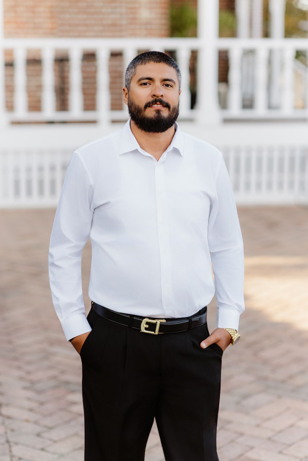 Bearded man in white dress shirt and black pants with gold belt buckle stands outdoors near white fence.