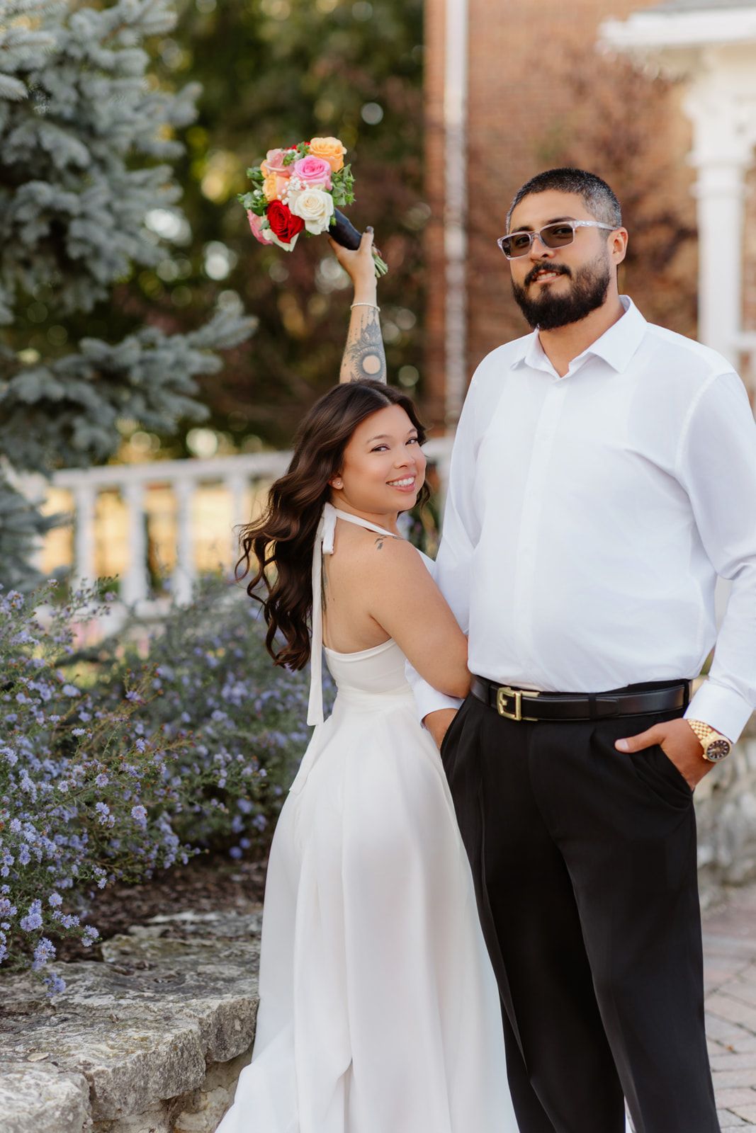 Bride in white halter gown raises colorful bouquet beside groom in white shirt and sunglasses outdoors.